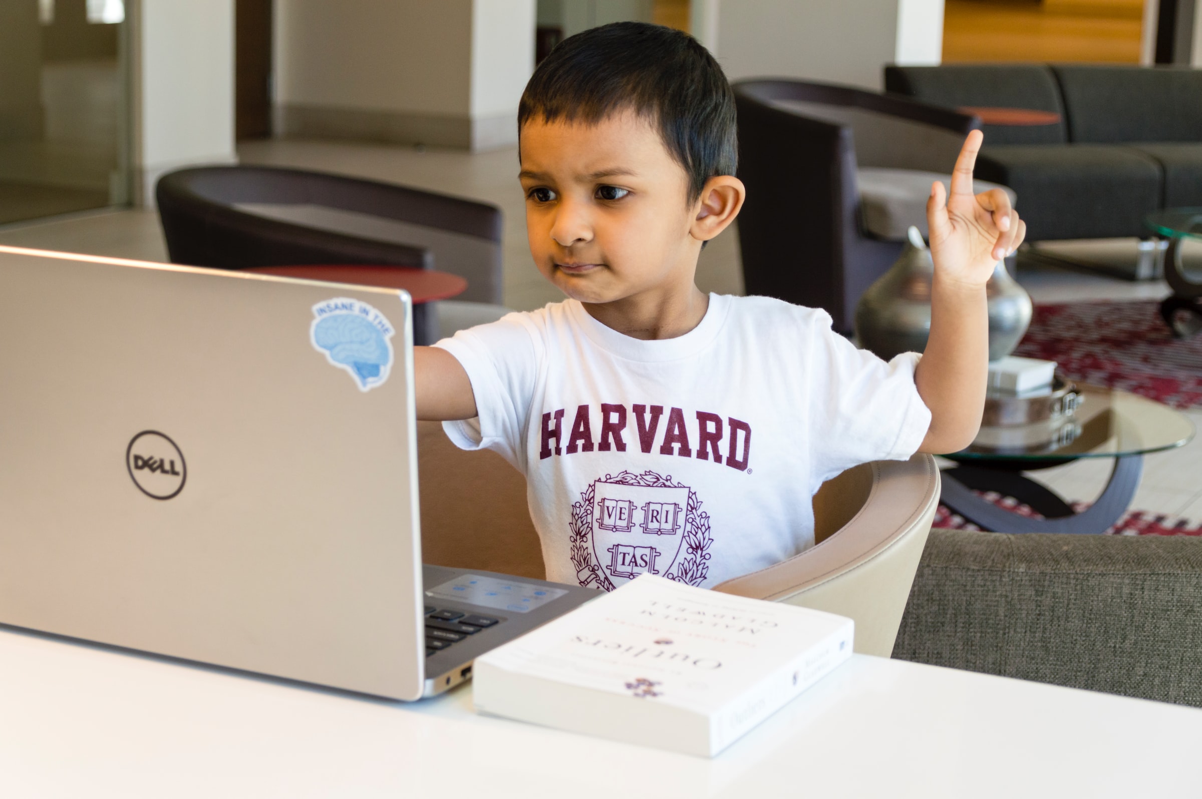 A child is wearing a white T-shirt, watching something in a gray Dell laptop, he is also raising his left hand.