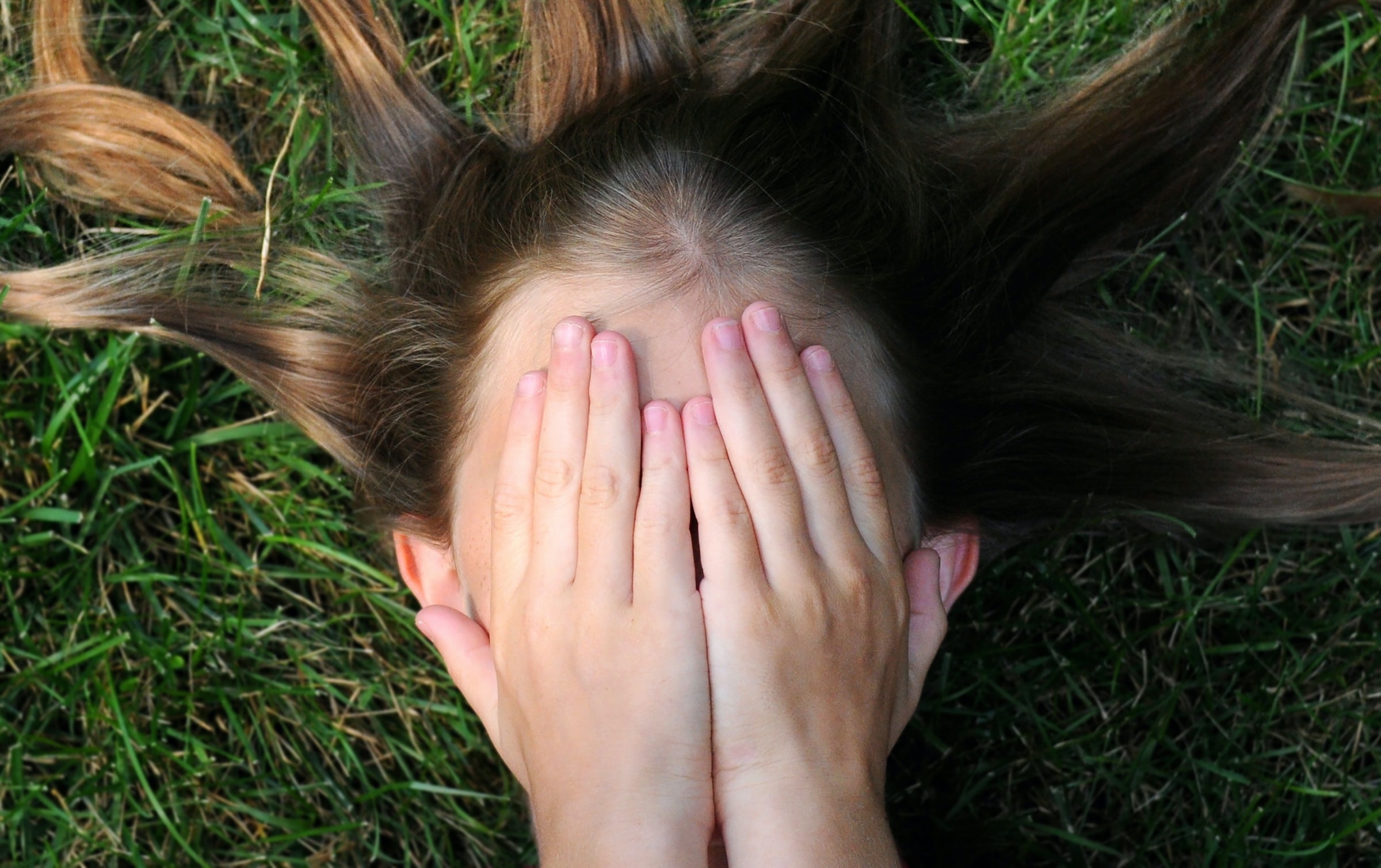 A girl is lying on the grass with her two hands on her face.