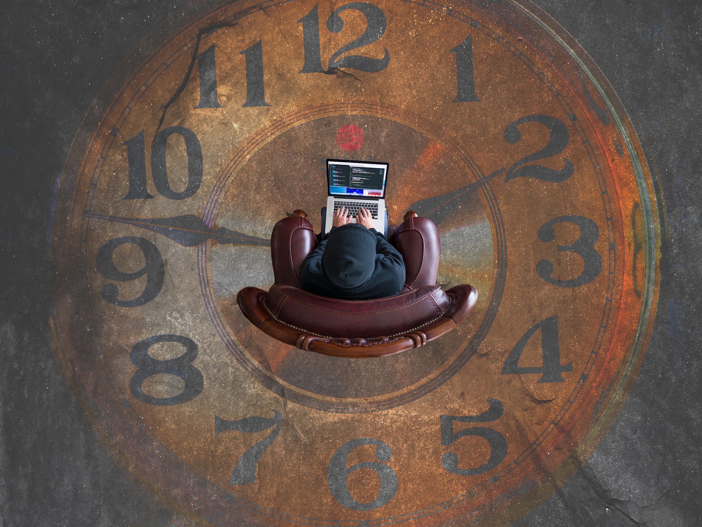 A huge clock with a man sitting in its center. The man is sitting in an armchair, he is typing something in a notebook.