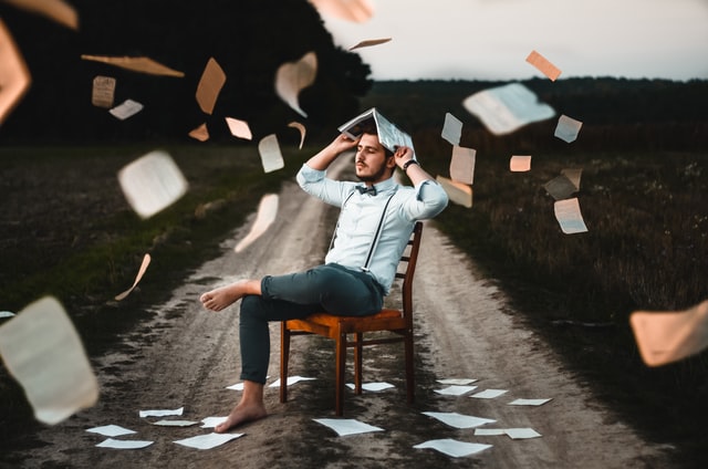 A man is sitting on a chair which is in the middle of a road, he has a book on his head and papers are falling around him.