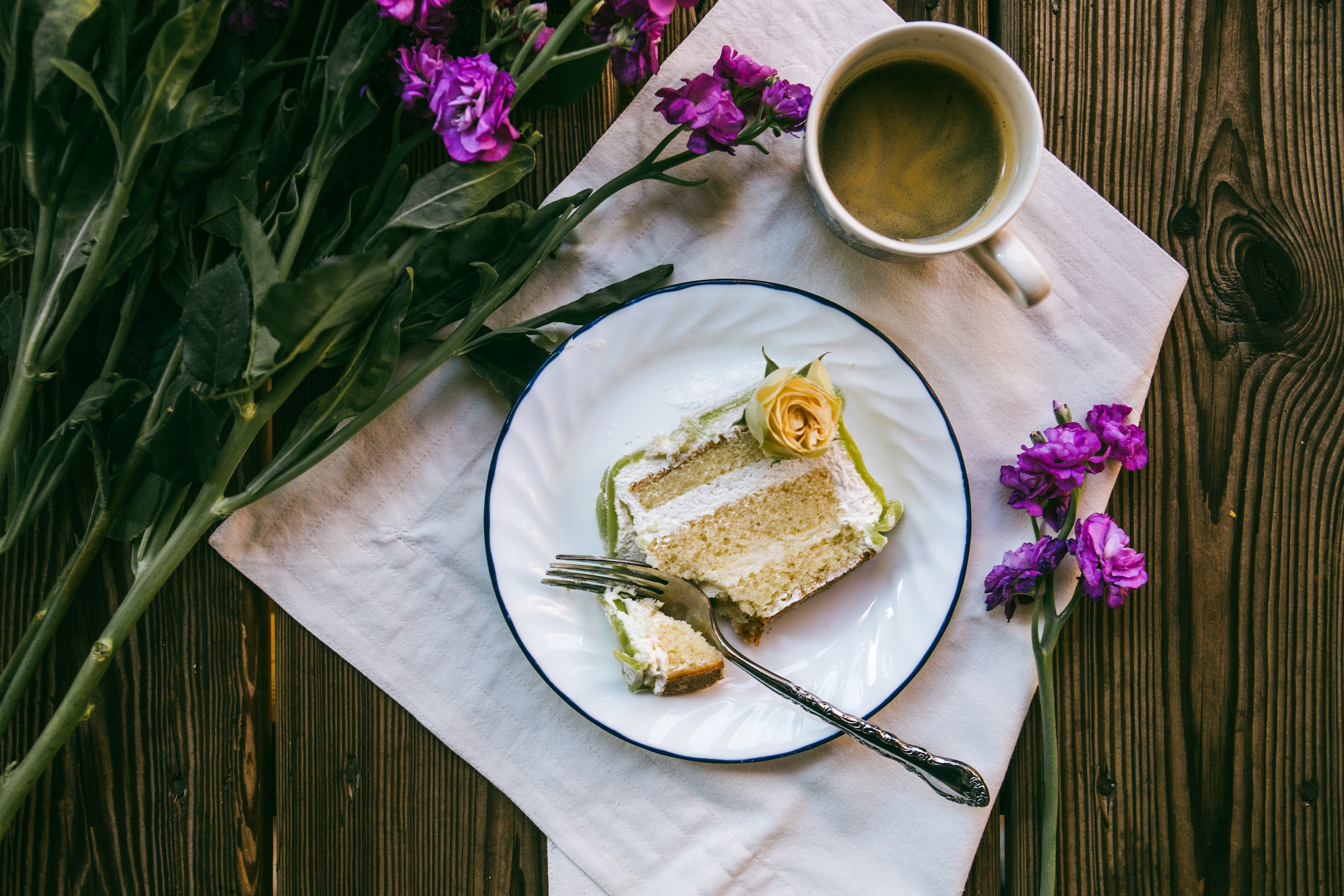 A creamy piece of cake is lying on a white plate. The plate is on a table, on the table there are also purple flowers and a cup with coffee.