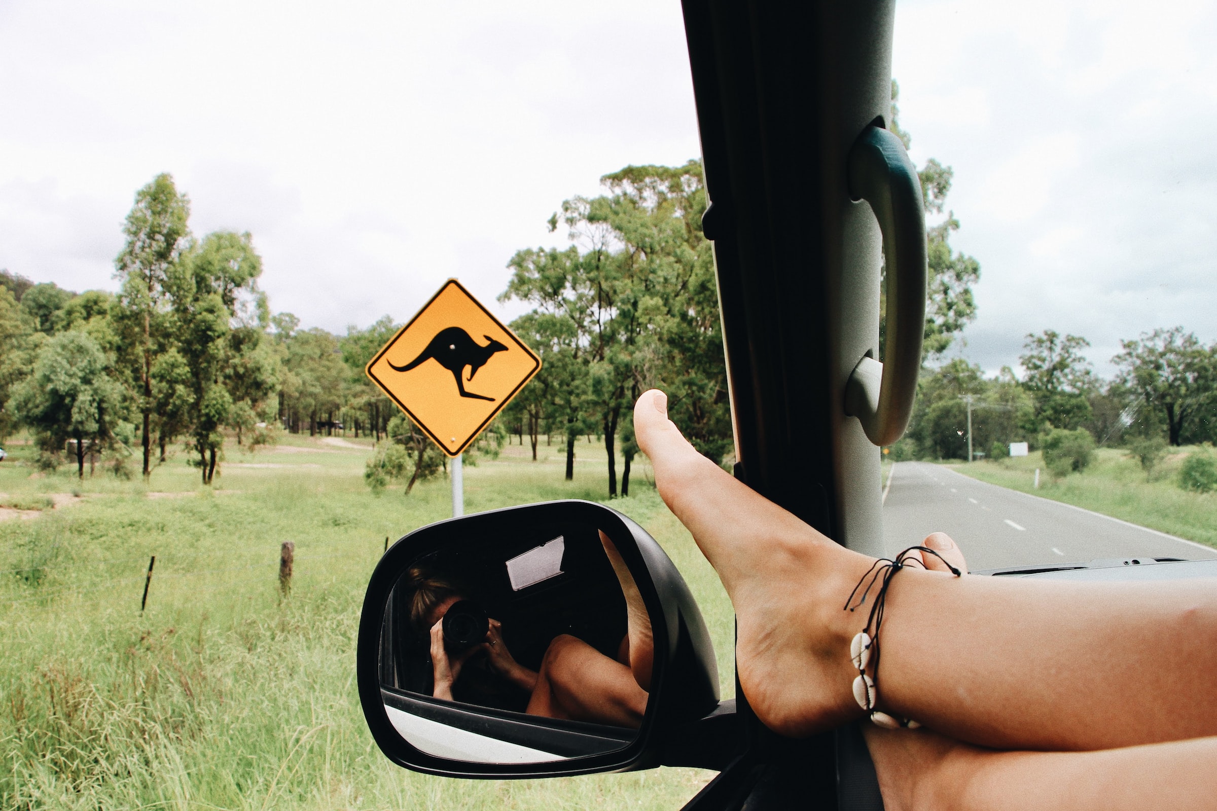 A car is on a road, inside of it there is a girl relaxed, who is taking a picture to a road kangaroo sign.