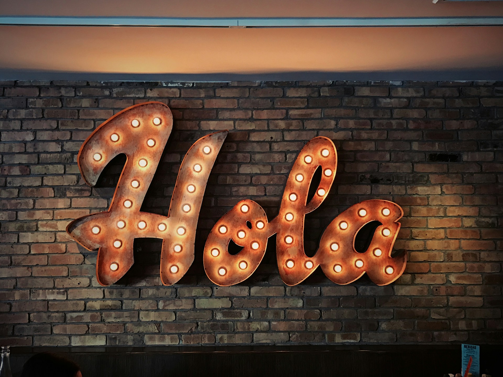 На кирпичной стене есть большая светящаяся вывеска, где написано “Hola”.