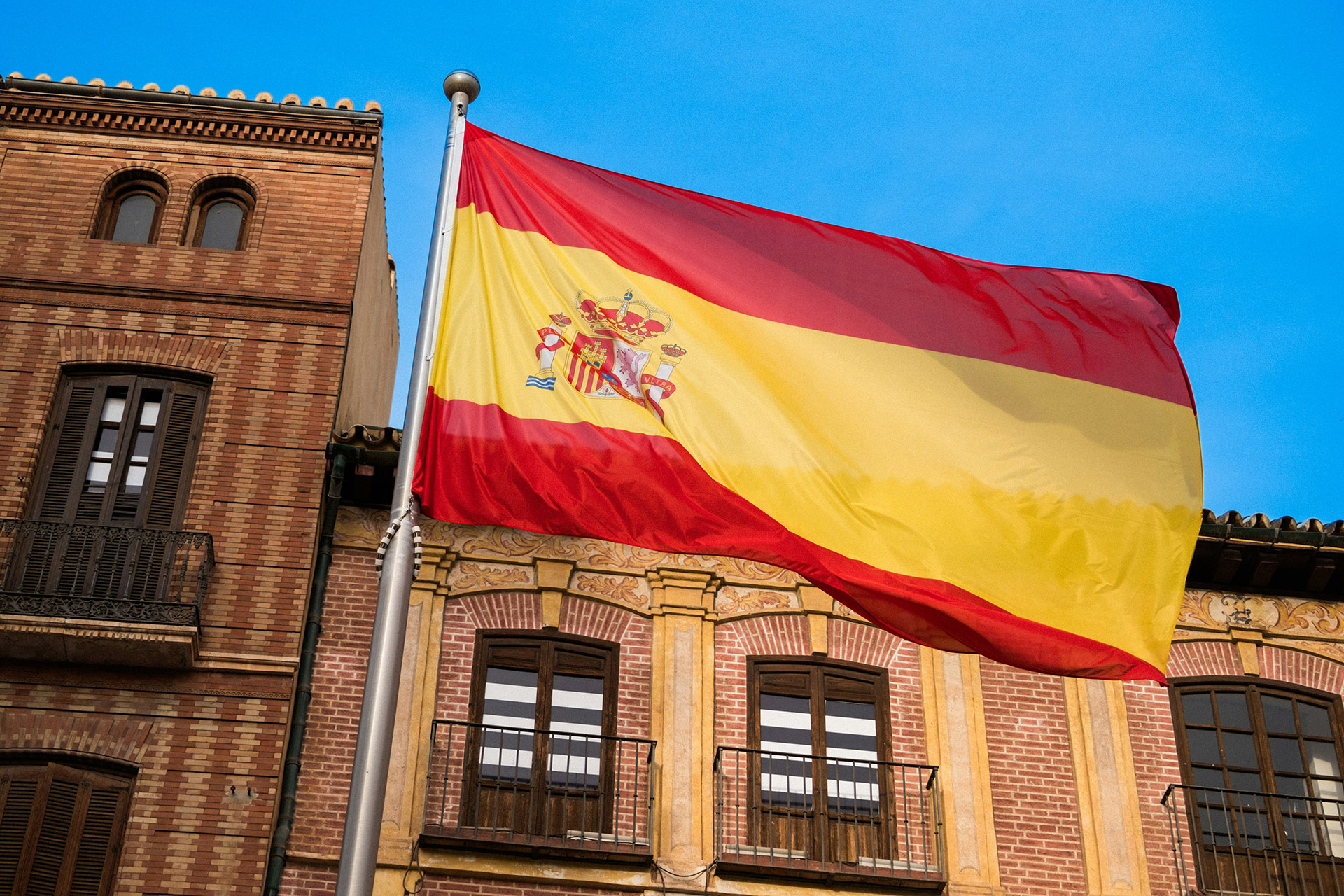 Флаг Испании реет на фоне старинного кирпичного здания и голубого неба.